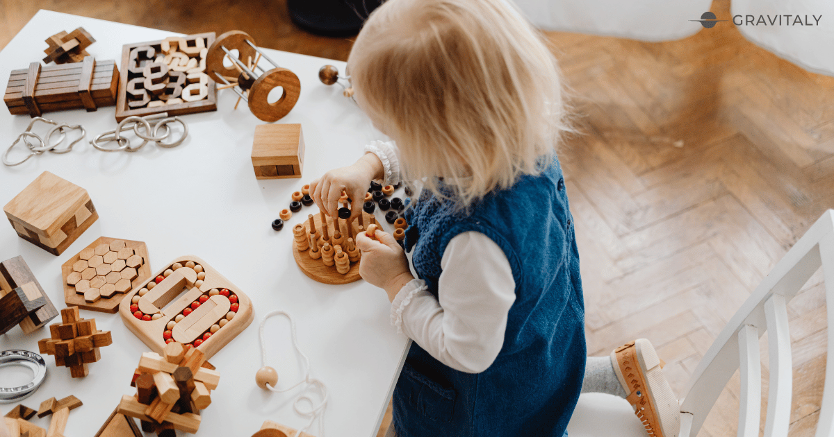 Top-down view of a toddler girl in a blue dress engaging in independent play with various natural wooden Montessori puzzles on a white table.
