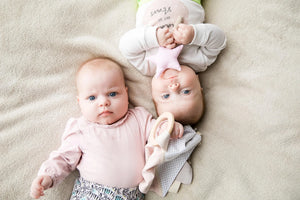 Dos bebés tumbados sobre una manta beige, cada uno con juguetes de peluche para bebé en colores pastel.