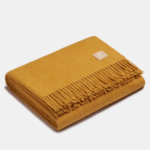 Coperta in lana di alpaca a spina di pesce color senape, piegata, con classiche frange finali e piccola etichetta quadrata ALPAKA in pelle, su sfondo chiaro.