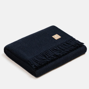 Coperta in lana di alpaca a spina di pesce blu navy, piegata, con classiche frange finali e piccola etichetta quadrata ALPAKA in pelle, su sfondo chiaro.