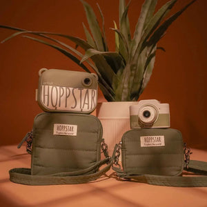 Hoppstar kids' cameras and olive green crossbody bags with 'Explore the world!' patches, styled with plant backdrop