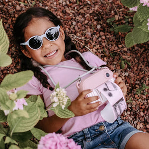Child holding pink instant camera with strap, wearing white sunglasses, lying on mulch near flowers
