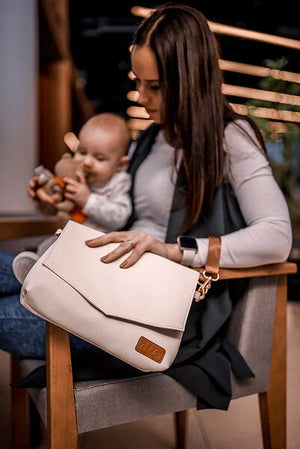 Lifestyle-Foto einer Mutter mit ihrem Baby, zeigt die elegante, hellcremefarbene Ally Cappuccino Kompakt-Wickeltasche mit kurzem braunen Handgelenksgurt.