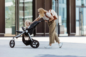 Foto lifestyle all'aperto di una donna bionda china su un passeggino nero a tre ruote per accarezzare il suo bambino. Indossa uno zaino in similpelle bianco e marrone.