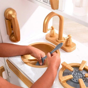 Gros plan sur les mains d'un enfant lavant des ustensiles jouets dans l'évier doré de la cuisine Angelove, mettant en valeur le faux plan de travail en marbre, le robinet doré et le téléphone mural.