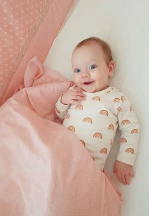 Bebé con body de estampado de arcoíris color crema, tumbado bajo una manta de lunares rosas.