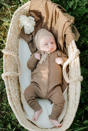 Baby im braunen Strickstrampler und Haube, liegt in einem Weidenkorb auf grünem Gras