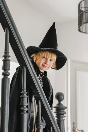 Child wearing black velour cape and witch hat beside a dark staircase