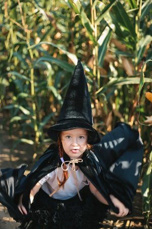 Child in black witch costume and magic cape holding the cape open in a corn field