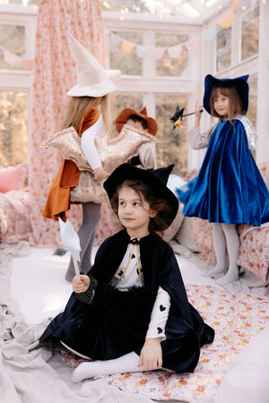 Child seated in black magic cape and witch hat with other fantasy-costumed children behind