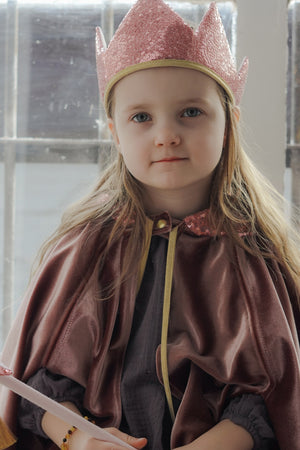 Close-up of a child wearing the rose velour Cosmic Pearl cape and sparkling crown beside a window