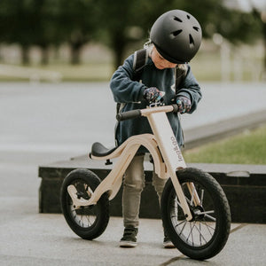 Child on natural wood balance bike with black tires and handlebars, wearing helmet outdoors