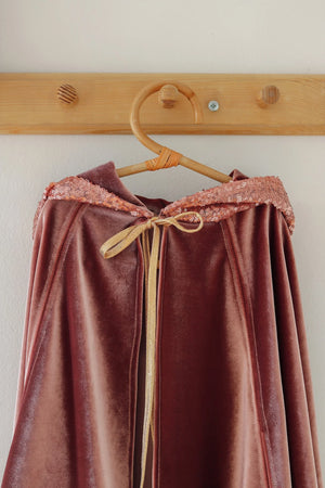 Pink velour Riding Hood cape hanging on a wooden hanger with sequin trim and gold tie fastening