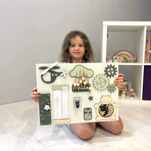 Child holding wooden activity board with green and cream gears, zippers, latches, and sensory elements for toddler development.