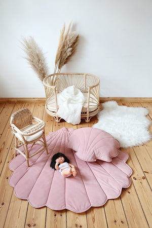 Doll resting on a powder pink shell playmat with a matching cushion and rattan nursery furniture