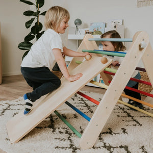 Wooden toddler climbing triangle with rainbow-colored rungs and slide, used by two children on patterned rug