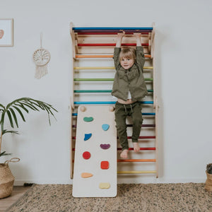 Child on wooden rainbow climbing ladder with colorful rungs and attached rock wall panel, natural wood finish
