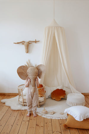 Child standing beneath a raw cotton canopy while holding a large rattan elephant mask