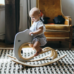 Baby on gray whale-shaped wooden rocker with natural wood base, nursery decor
