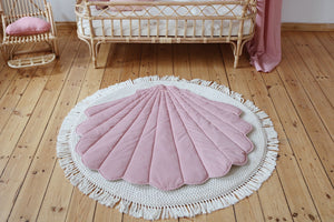 Powder pink shell playmat centred on a round fringed rug beside a rattan children’s bed