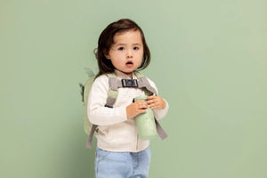 Tout-petit tenant une gourde inox verte motif dragon, portant un pull beige et un sac à dos.