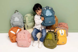 Child sitting among colourful Trixie animal backpacks while holding a blue dinosaur backpack