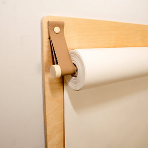 Light wood drawing board with tan leather strap holding white paper roll, minimalist nursery art station