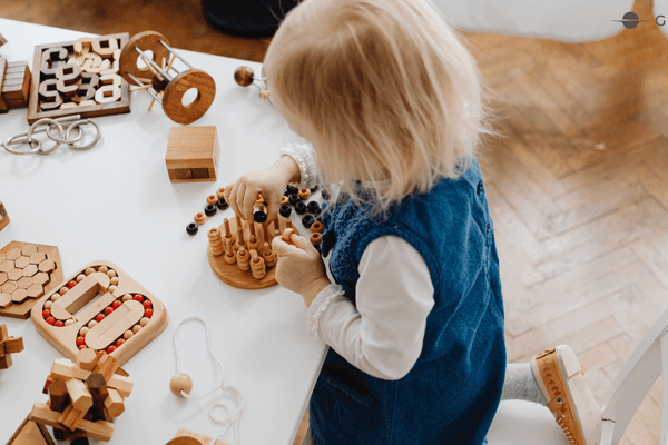 Top-down view of a toddler girl in a blue dress engaging in independent play with various natural wooden Montessori puzzles on a white table.
