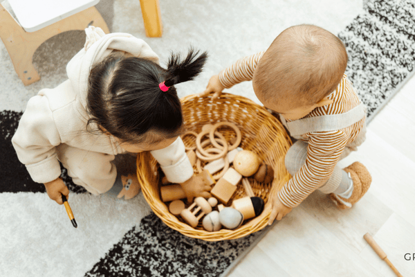 Two young children sitting on a soft rug, collaboratively playing with natural wooden Montessori toys in a sunlit, minimalist nursery.