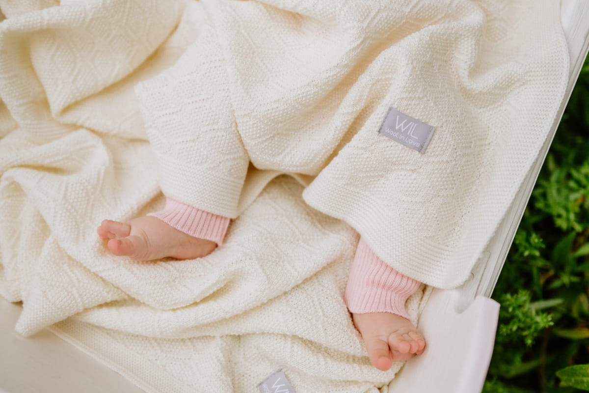 Dettaglio di piedini da bebè in calzine di maglia rosa, avvolti in una soffice coperta color crema a rombi. La texture evoca calore, comfort e lusso delicato.