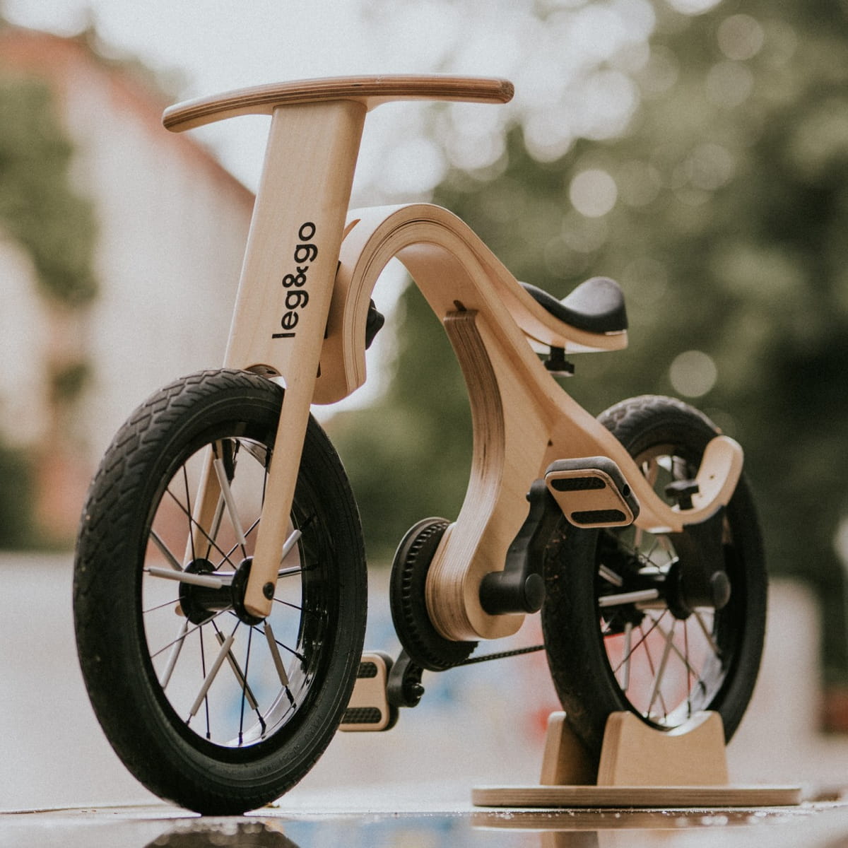 Vista laterale della bici in legno 3-in-1 Leg&Go con kit pedali: telaio in betulla naturale, pneumatici neri e trasmissione a cinghia su sfondo outdoor sfocato.