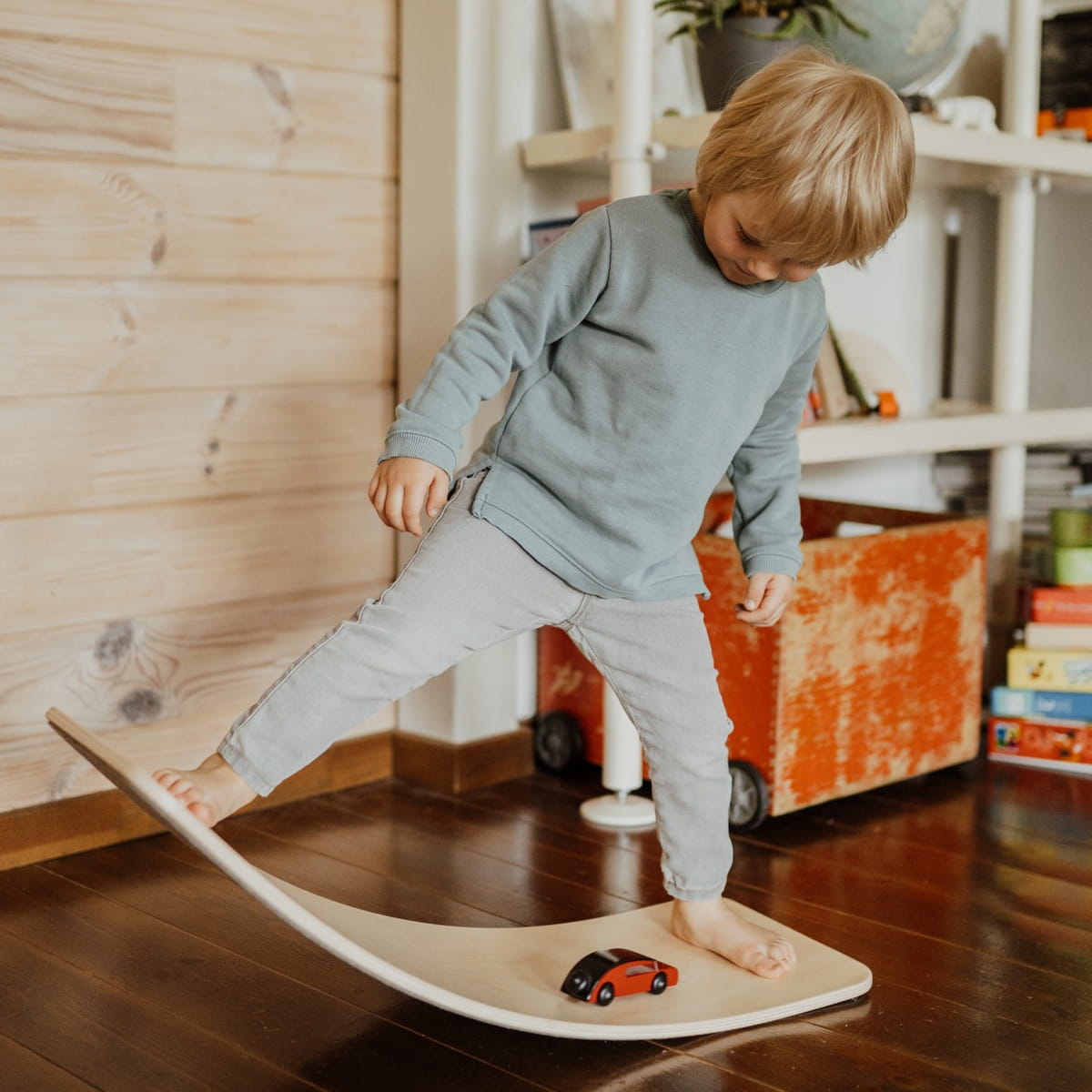 A side view of a Leg&Go curved wooden balance board with a natural wood finish, showing a black metal support structure and large black wheels, on a blurred background