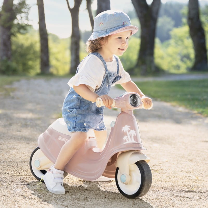 Un bambino con salopette in denim e cappellino cavalca un monopattino vintage rosa con ruote bianche lungo un sentiero sterrato, sorridente.