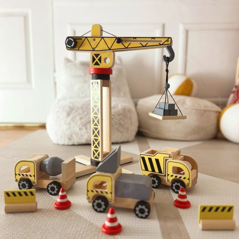 Set di costruzioni in legno con gru gialla che solleva un pallet, betoniera, camion con cassone ribaltabile e coni stradali su un tappeto gioco.