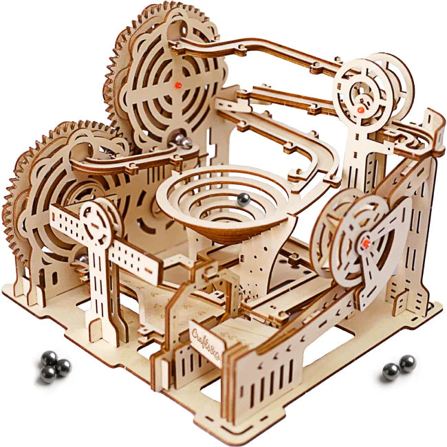 Intricato puzzle meccanico 3D in legno (pista per biglie) con ingranaggi tagliati al laser, piste a chiocciola, imbuto decorato e biglie in argento su fondo bianco.