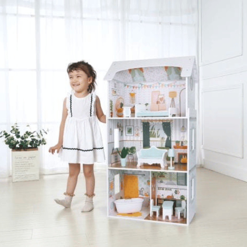 A happy young girl stands and smiles next to a four-story wooden dollhouse, which is completely open to reveal distinct levels with detailed miniature furniture and wallpapers, set on a light wood floor in a bright room.