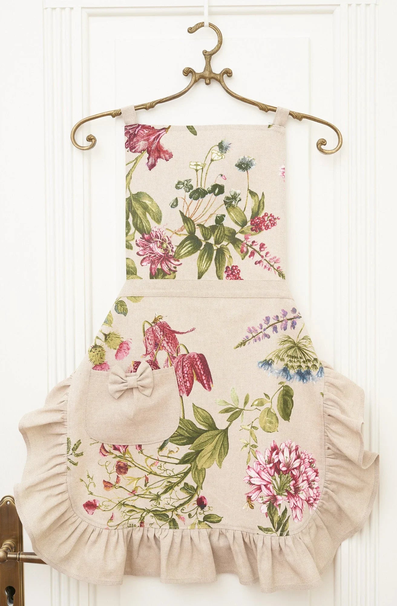 Floral print apron with ruffles and bow pocket, hanging on brass hanger against white door