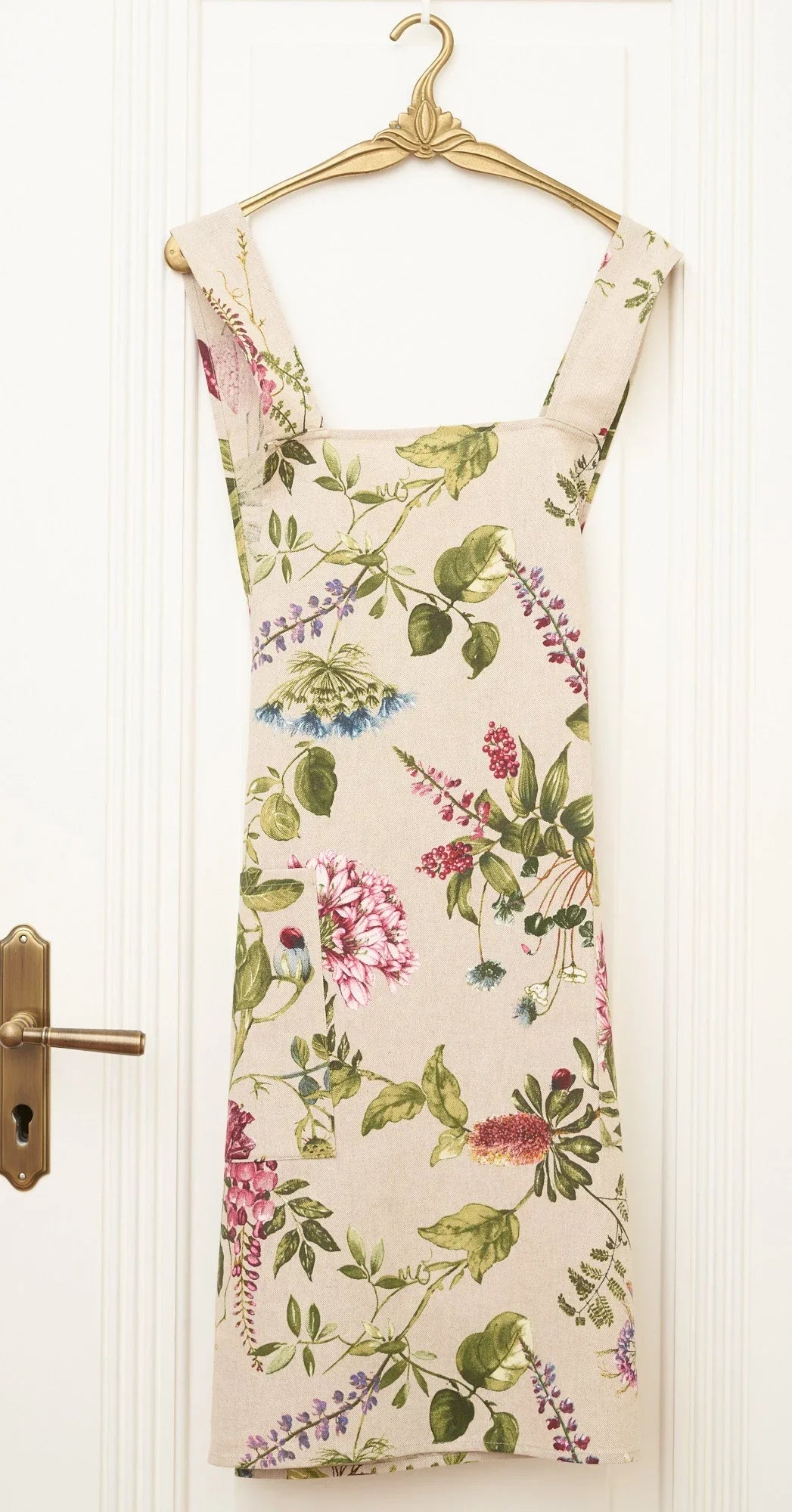 Floral print linen apron with green leaves and pink purple flowers, hanging on gold hanger against white door