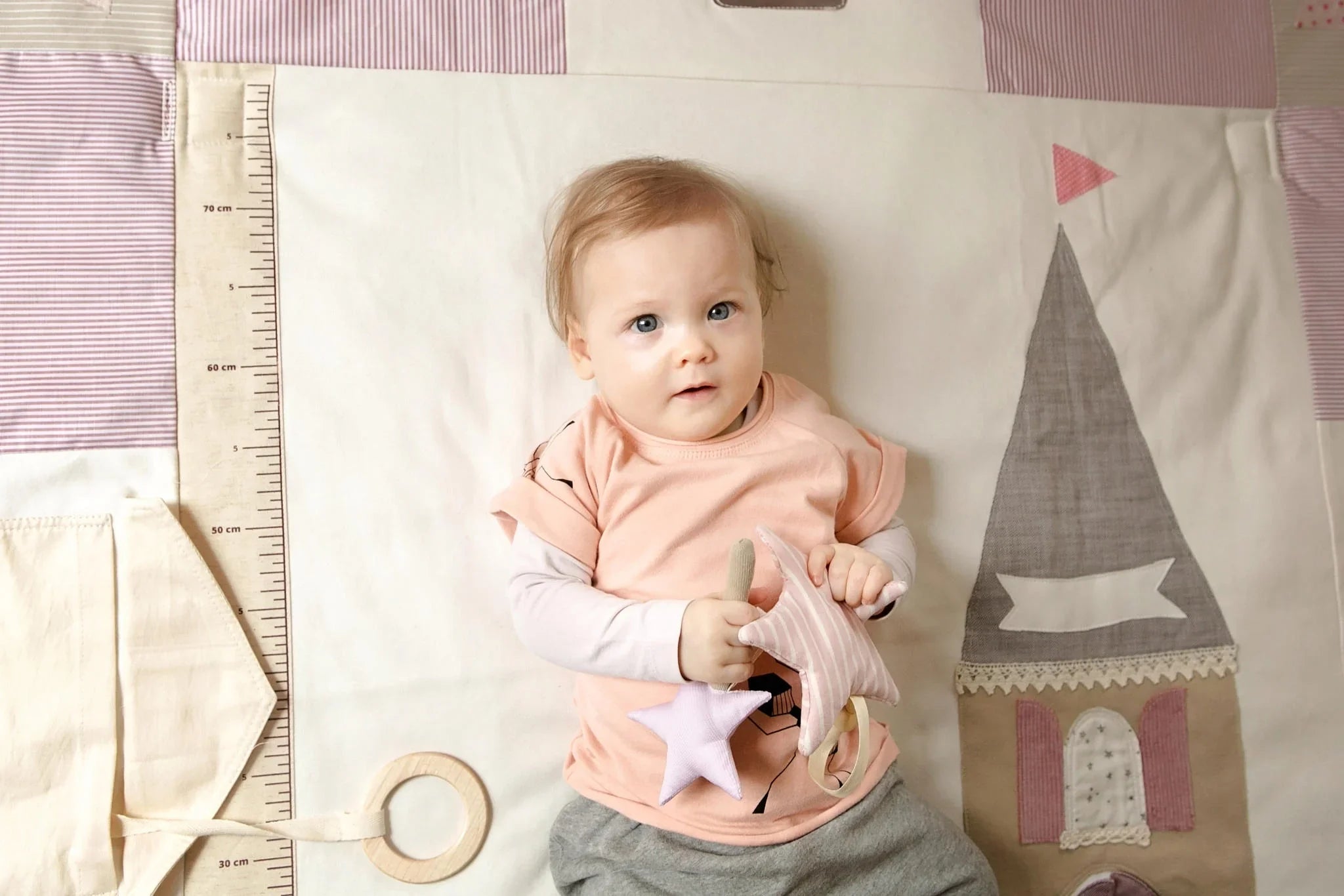 Baby on soft nursery play mat with growth chart and pastel castle appliqué, holding plush star toys