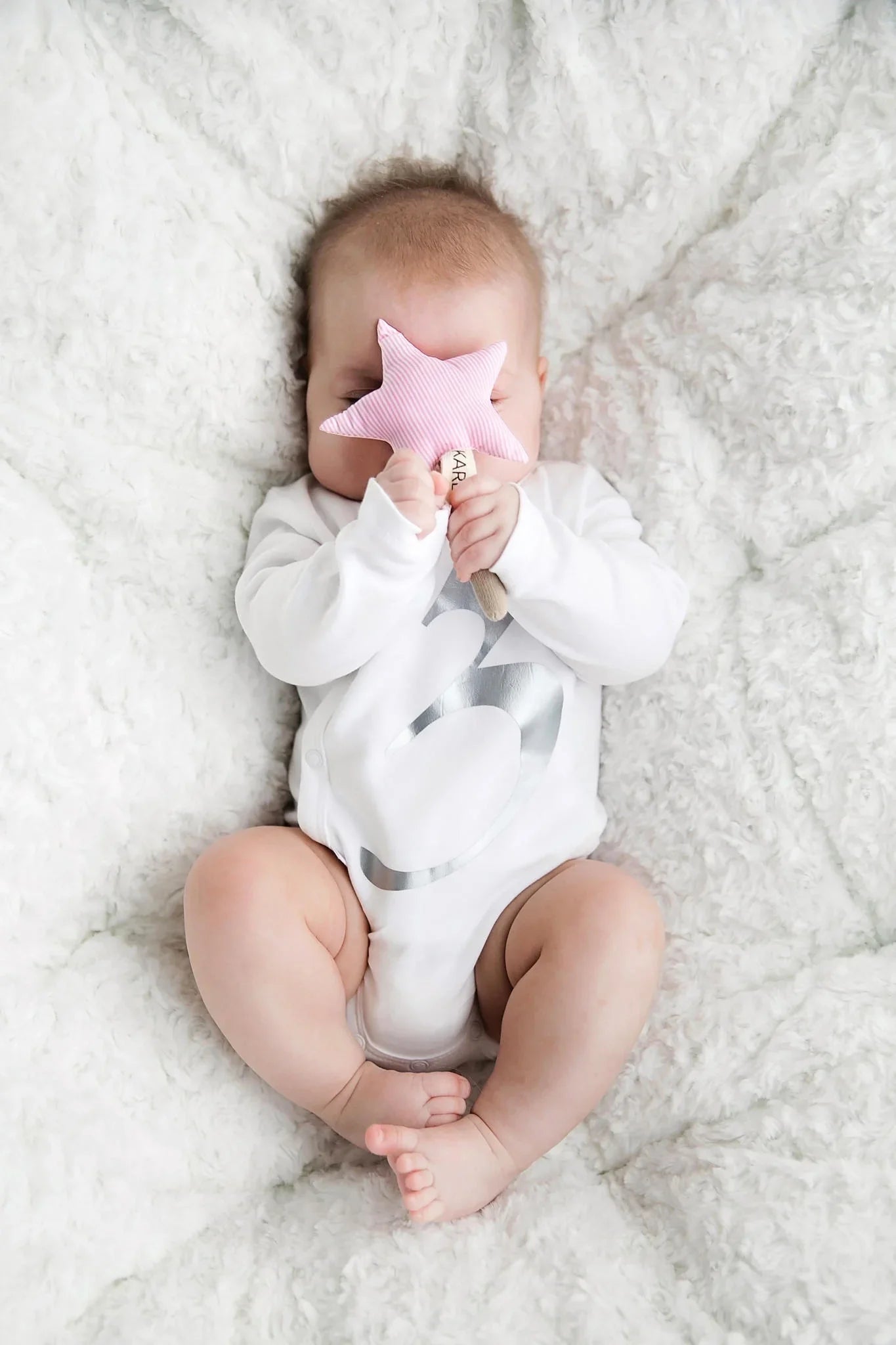 Bebé con body blanco sosteniendo sonajero de peluche de estrella rosa sobre manta blanca de textura suave.