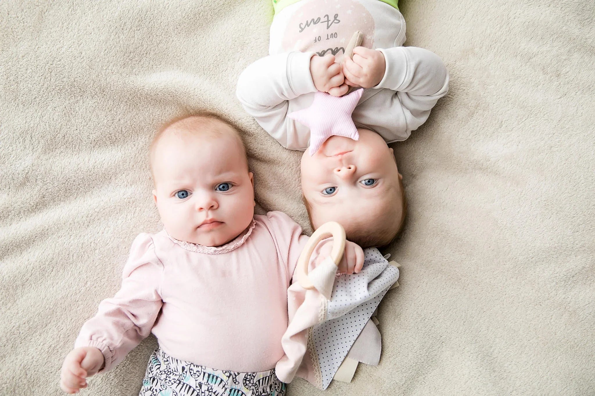 Dos bebés tumbados sobre una manta beige, cada uno con juguetes de peluche para bebé en colores pastel.