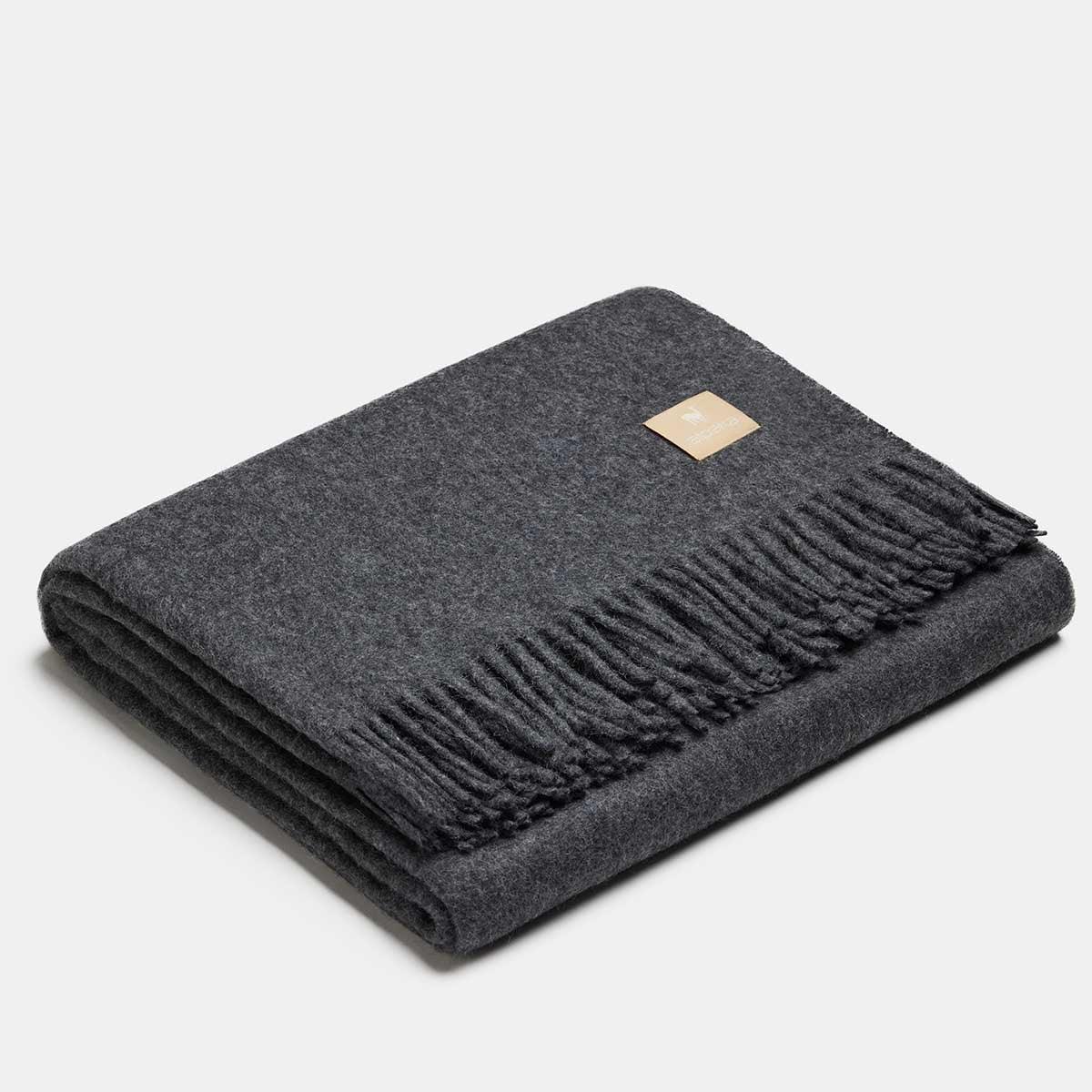 Couverture gris foncé pliée avec étiquette dorée sur fond gris clair. Laine Alpaka.