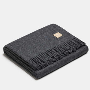 Couverture gris foncé pliée avec étiquette dorée sur fond gris clair. Laine Alpaka.