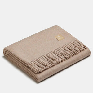 Couverture jetée en laine d'alpaga café au lait à motif chevrons, pliée avec soin, finitions à franges classiques et petit écusson en cuir ALPAKA sur fond clair