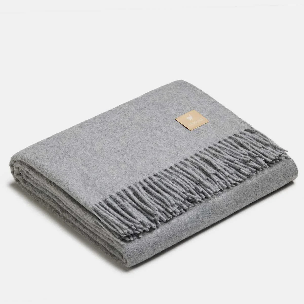 Couverture jetée en laine d'alpaga argent à motif chevrons, pliée avec soin, finitions à franges classiques et petit écusson en cuir ALPAKA sur fond clair.