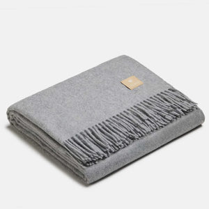 Couverture jetée en laine d'alpaga argent à motif chevrons, pliée avec soin, finitions à franges classiques et petit écusson en cuir ALPAKA sur fond clair.