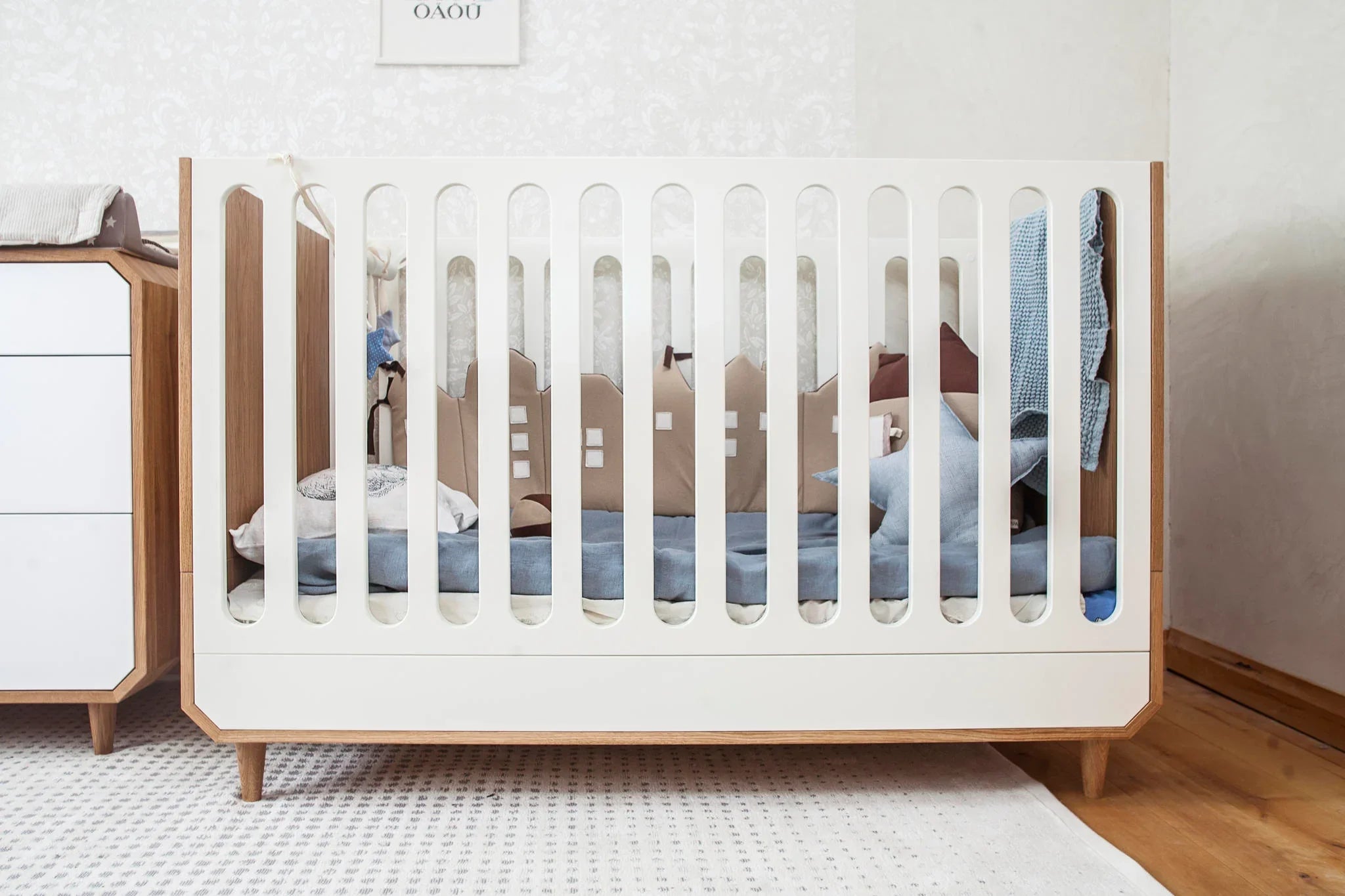 Modern wooden baby crib with white slats, blue bedding, and decorative cushions in a nursery