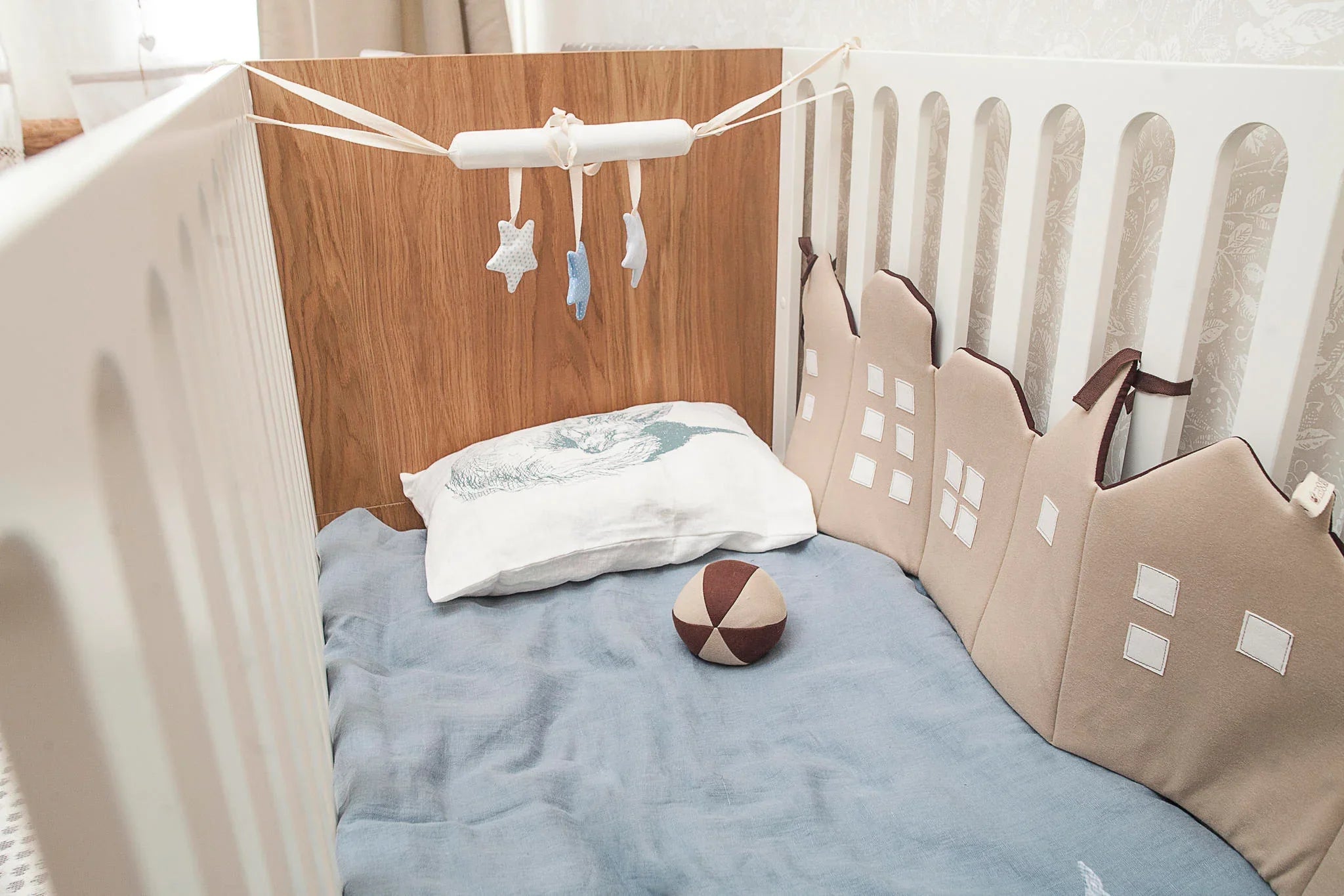 Modern baby crib with neutral bedding, house-shaped crib bumper, fabric ball, and star mobile