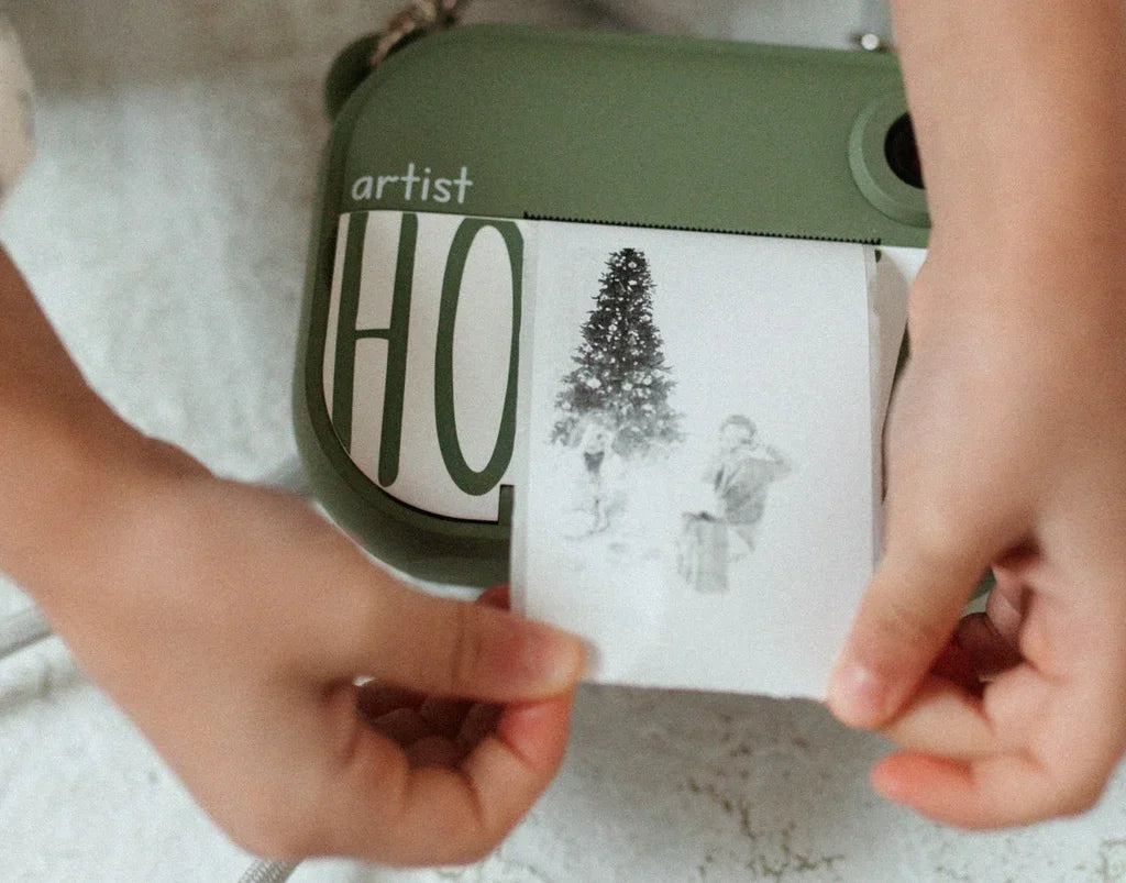 Olivgrüne Sofortbildkamera beim Drucken eines Fotos von einem Weihnachtsbaum und Kindern, beschriftet als „Artist“