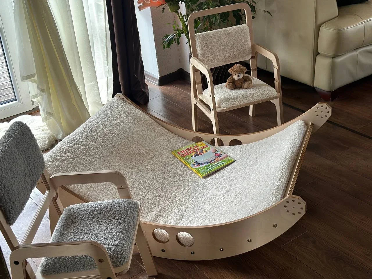Wooden toddler rocking chair with cream shearling cushion, paired with matching small chair and gray cushion, in cozy nursery setting.